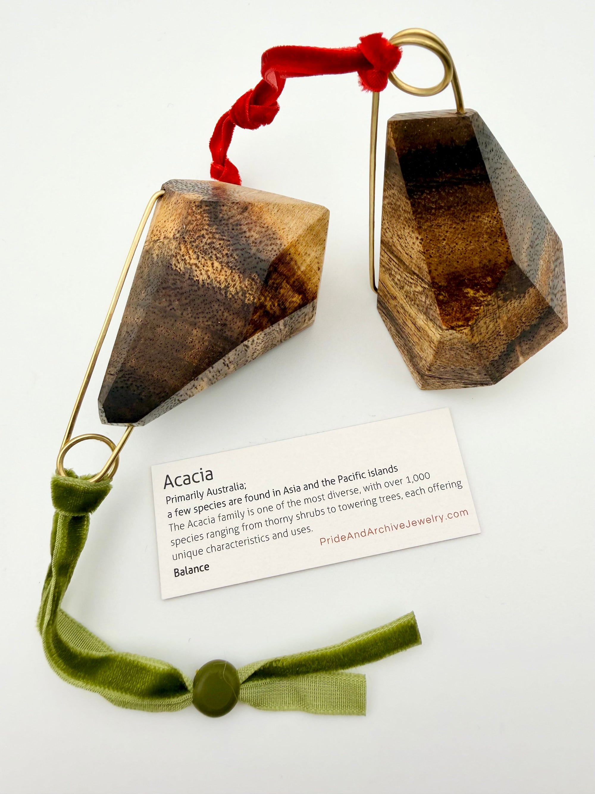 Pair of wood geometric ornaments with brass details and red and green ribbons on a white background and tag that talks about Acacia wood facts