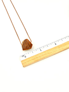 Blonde wood pendant with a dramatic grain pattern on rose gold ball chain next to ruler for scale on a white background
