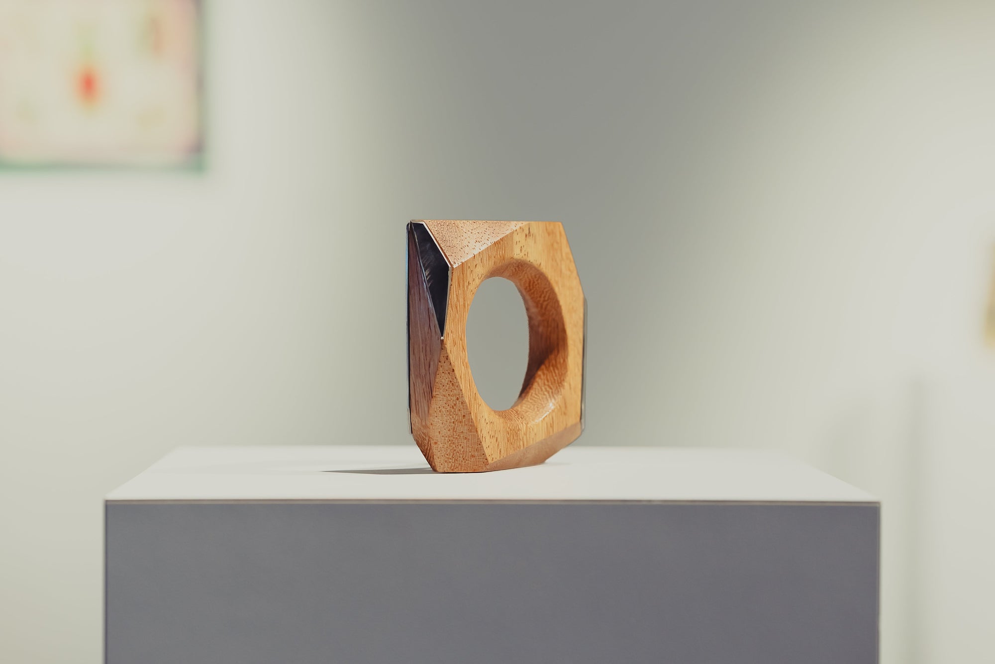 Wooden geometric ring sculpture on a white pedestal with a blurred art gallery in the background