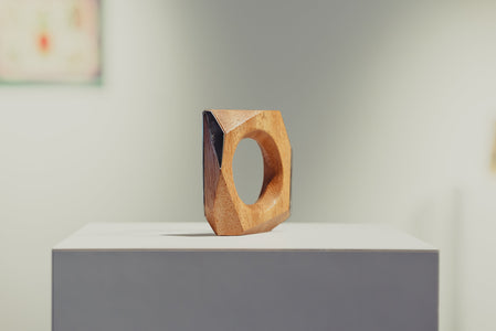 Wooden geometric ring sculpture on a white pedestal with a blurred art gallery in the background