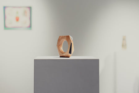 Wooden geometric ring sculpture on a white pedestal with a blurred art gallery in the background
