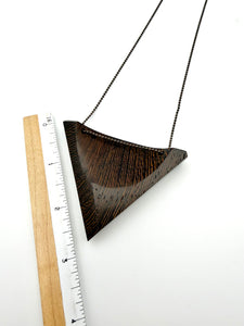 Triangular wooden pendant with oxidized chain on a white background with a ruler for scale.
