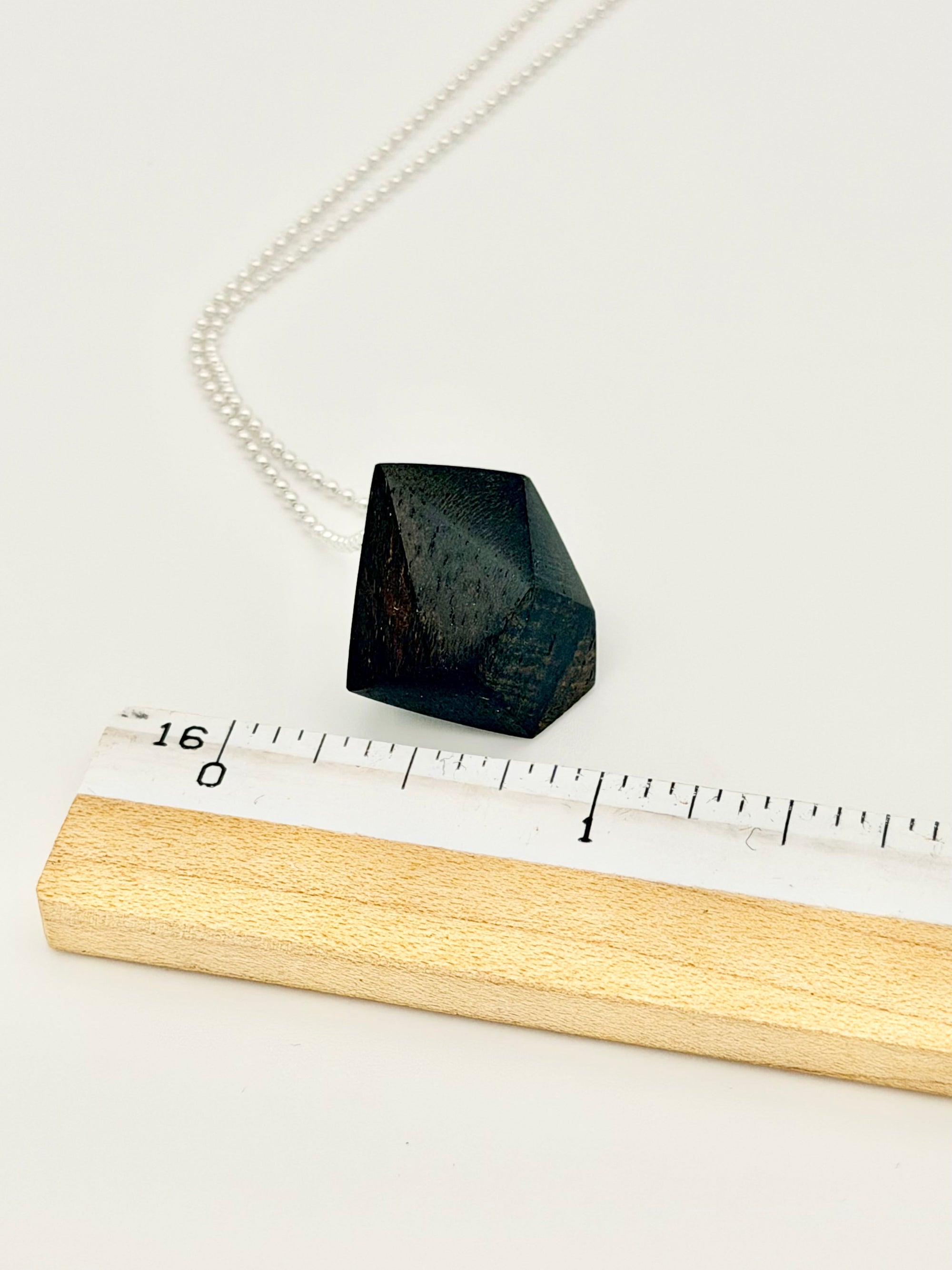 Black Geometric wood pendant on silver chain next to ruler for scale. 